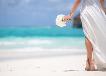 A bride walking barefoot on the sand whilst carrying a small bouquet of flowers