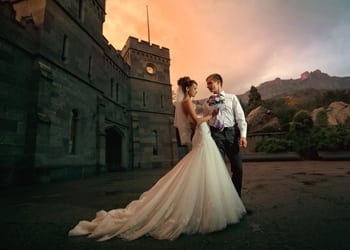 A bride and groom posing in front of a castle during sunset